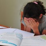 Criança esconde o rosto nas mãos. Na frente dela, em cima da mesa, há livros e cadernos